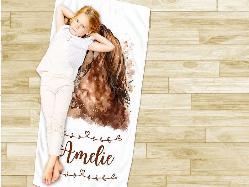 May include: White beach towel with a watercolor painting of a brown horse's head and a girl lying on the towel. The towel has the name "Amelie" written in brown cursive with decorative hearts.