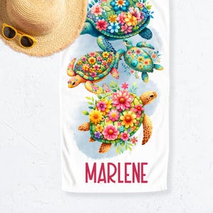 Könnte beinhalten: Weißes Strandtuch mit einem Design von drei Meeresschildkröten, die mit bunten Blumen geschmückt sind. Die Schildkröten sind in Grün-, Gelb- und Orangetönen gehalten. Der Name "MARLENE" ist unten rot aufgedruckt. Ein Strohhut und eine gelbe Sonnenbrille befinden sich in der oberen linken Ecke.