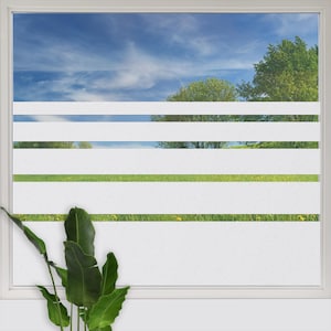 Könnte beinhalten: Eine Fensterscheibe mit einer dekorativen Sichtschutzfolie, die vier horizontale, mattweiße Streifen aufweist. Die Folie erzeugt einen halbtransparenten Effekt und verdeckt teilweise den Blick auf einen blauen Himmel, grüne Bäume und eine Wiese.