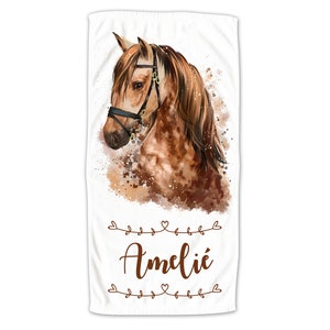 May include: A white beach towel with a watercolor illustration of a brown and white horse's head. The horse is wearing a black bridle with gold accents. The towel has the name "Amelie" written in brown cursive lettering.