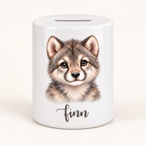 Könnte beinhalten: Eine weiße, zylindrische Spardose aus Keramik mit einer detaillierten Illustration eines flauschigen Wolfswelpen mit braunem und grauem Fell. Der Name Finn ist in einer dunkelgrauen Schreibschrift unter dem Tierporträt aufgedruckt.
