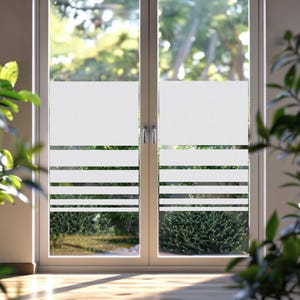 Könnte beinhalten: Eine weiße Fensterfolie, die auf einem Doppelfenster angebracht ist. Die Folie hat eine mattierte Oberseite und horizontale weiße Streifen, die einen Sichtschutz bilden. Das Fenster ist in einem weißen Rahmen, mit Grün draußen sichtbar.