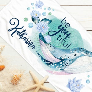 Könnte beinhalten: Ein weißes Strandtuch mit einer Aquarell-Illustration eines blauen Wals mit rosa Unterleib, umgeben von blauem und grünem Wasser und weißen Blumen. Der Text "be you tiful" ist in blauer Kursivschrift geschrieben und der Name "Katharina" ist in blauer Kursivschrift geschrieben.