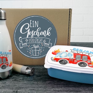 Könnte beinhalten: Ein Geschenkset mit einem weißen und roten Feuerwehrauto-Design. Das Set enthält eine Lunchbox, eine Wasserflasche und einen Strohhalm. Die Lunchbox und die Wasserflasche sind mit dem Namen "Benjamin" versehen. Die Box hat einen Aufkleber mit der Aufschrift "Ein Geschenk fur dich!"