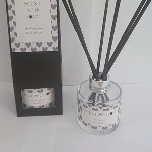 May include: A black box with a white label that says "SCENT WITH LOVE Sparkling Lemon Reed Diffuser" and a heart pattern. The box is next to a clear glass bottle with a silver lid and black sticks. The bottle has a white label that says "SCENT WITH LOVE Sparkling Lemon Reed Diffuser" and a heart pattern.