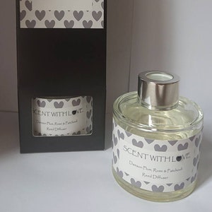 May include: A clear glass reed diffuser with a silver cap and a matching box. Both the diffuser and box feature a label with the text "SCENT WITH LOVE" and the scent description "Damson Plum, Rose & Patchouli". The labels are decorated with grey hearts.