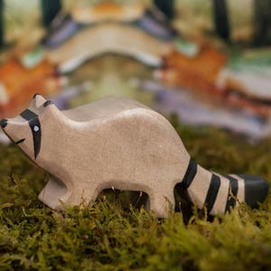 May include: A wooden raccoon figurine with a black and white striped tail. The figurine is painted in a light brown color and has a realistic appearance. The raccoon is standing on a bed of green moss.