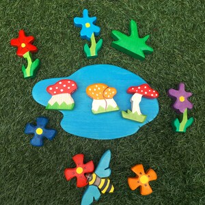 May include: A wooden puzzle set with a blue pond, red, orange, and white mushrooms, green leaves, and purple, blue, and red flowers.