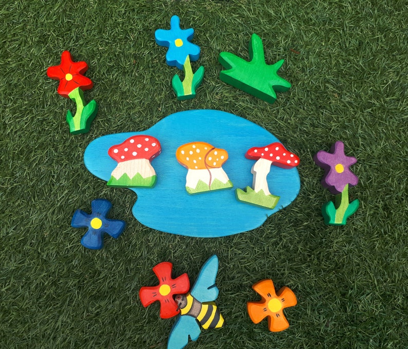 May include: A wooden puzzle set with a blue pond, red, orange, and white mushrooms, green leaves, and purple, blue, and red flowers.
