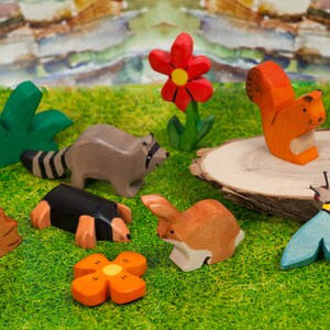 May include: Wooden toy animals and flowers on a green surface. The animals include a raccoon, a squirrel, a hedgehog, a mole, a rabbit, and a bee. The flowers are red and orange.