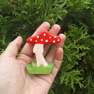 May include: A wooden mushroom toy with a red cap with white dots and a green base. The mushroom is held in a hand.