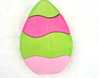 May include: A wooden egg-shaped puzzle toy with three pieces. The top piece is green, the middle piece is pink, and the bottom piece is green.