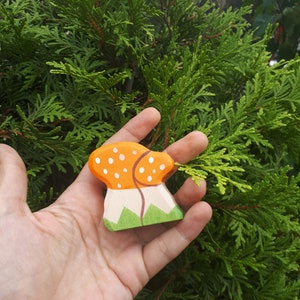 May include: A wooden toy mushroom with a white stem and orange cap with white dots. The mushroom is painted with green grass at the base.