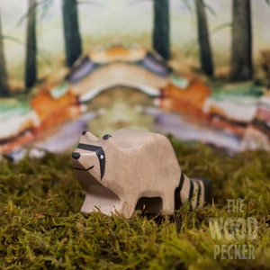 May include: A wooden raccoon figurine with black eyes and a black stripe across its eyes. The figurine is standing on a bed of green moss with a blurred background of trees.