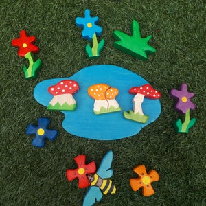 May include: A wooden toy set with a blue pond, mushrooms, flowers, and a bee. The flowers are red, blue, purple, and orange. The mushrooms are red and white, orange and white, and red. The bee is black and yellow.