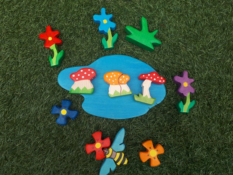 May include: A wooden toy set with a blue pond, mushrooms, flowers, and a bee. The flowers are red, blue, purple, and orange. The mushrooms are red and white, orange and white, and red. The bee is black and yellow.