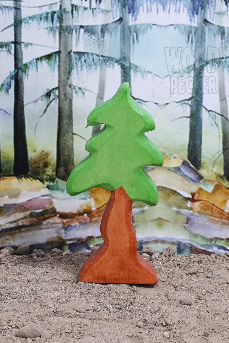 May include: A wooden toy tree with a green top and a brown trunk. The tree is standing on a sandy surface in front of a watercolor background of trees.