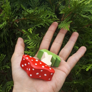 May include: A hand holds two wooden toy mushrooms. One mushroom is red with white polka dots and the other is green.