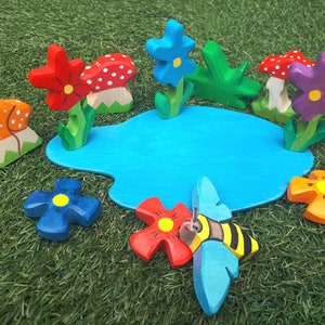 May include: A wooden toy set featuring a blue pond, red, orange, and purple flowers, a bee, and mushrooms with white polka dots.