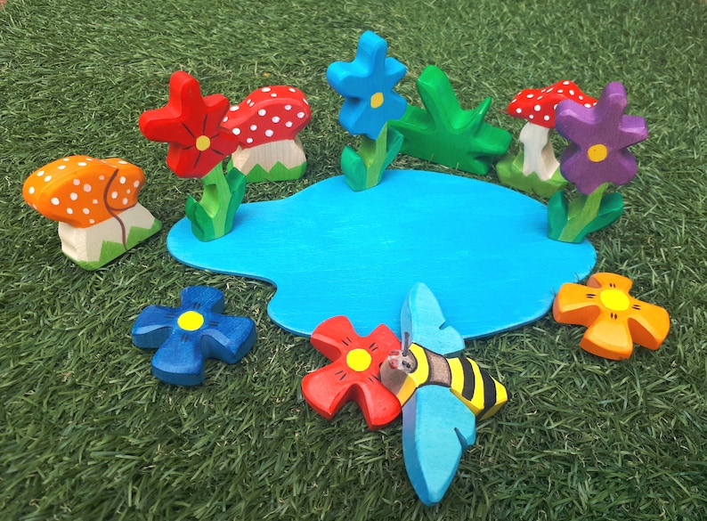 May include: A wooden toy set featuring a blue pond, red, orange, and purple flowers, a bee, and mushrooms with white polka dots.
