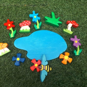 May include: A wooden toy set featuring a blue pond, red, orange, and yellow mushrooms, purple, blue, and red flowers, and a green bush. The set also includes a bee with black and yellow stripes.