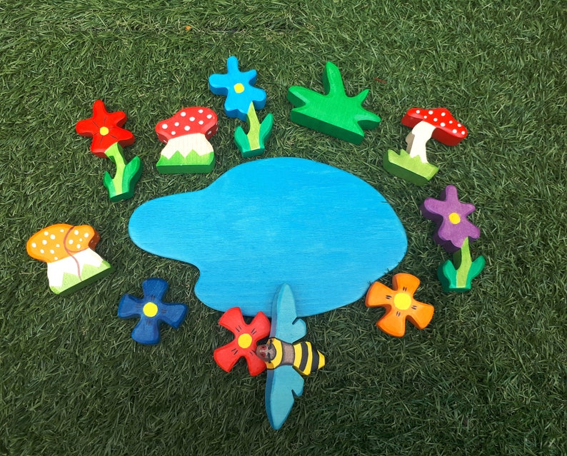 May include: A wooden toy set featuring a blue pond, red, orange, and yellow mushrooms, purple, blue, and red flowers, and a green bush. The set also includes a bee with black and yellow stripes.