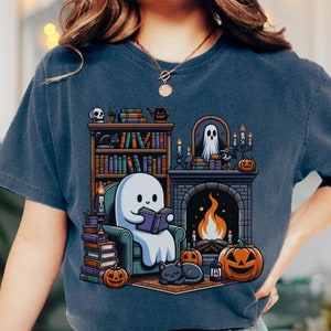 May include: A navy blue t-shirt featuring a cartoon ghost reading a book in front of a fireplace, with pumpkins and a black cat. A bookshelf and candles are also in the design. Halloween themed.