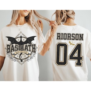 Pode incluir: T-shirts brancas com estampas gráficas. Uma camiseta apresenta "BASGIATH WAR COLLEGE" com um dragão e uma faixa. A outra mostra "RIORSON 04" com um pequeno gráfico de pássaro. Ambas as camisetas têm detalhes em preto e dourado.