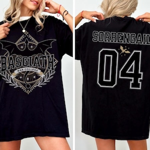 Black oversized t-shirt inspired by Fourth Wing featuring a bold ‘Basgiath War College’ crest on the front with swords, wings, stars, and laurel branches. The back showcases the name ‘Sorrengail’ above the number ‘04’ in a varsity-style font, with a small dragon graphic above the numbers. Modeled by a blonde woman, showing both front and back views. Perfect for fans of the Riders Quadrant and fantasy academy themes