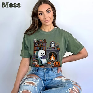 May include: Moss green t-shirt featuring a cartoon ghost reading a book in front of a fireplace, surrounded by pumpkins, books, and a black cat. The word "Moss" is printed in the upper left corner. The shirt is worn by a person wearing blue jeans.