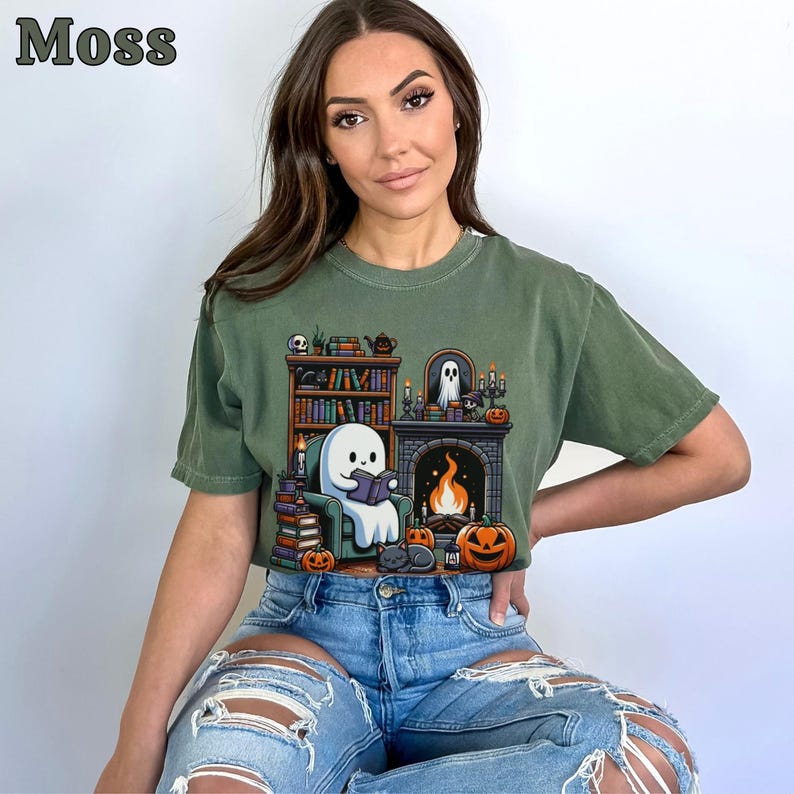 May include: Moss green t-shirt featuring a cartoon ghost reading a book in front of a fireplace, surrounded by pumpkins, books, and a black cat. The word "Moss" is printed in the upper left corner. The shirt is worn by a person wearing blue jeans.