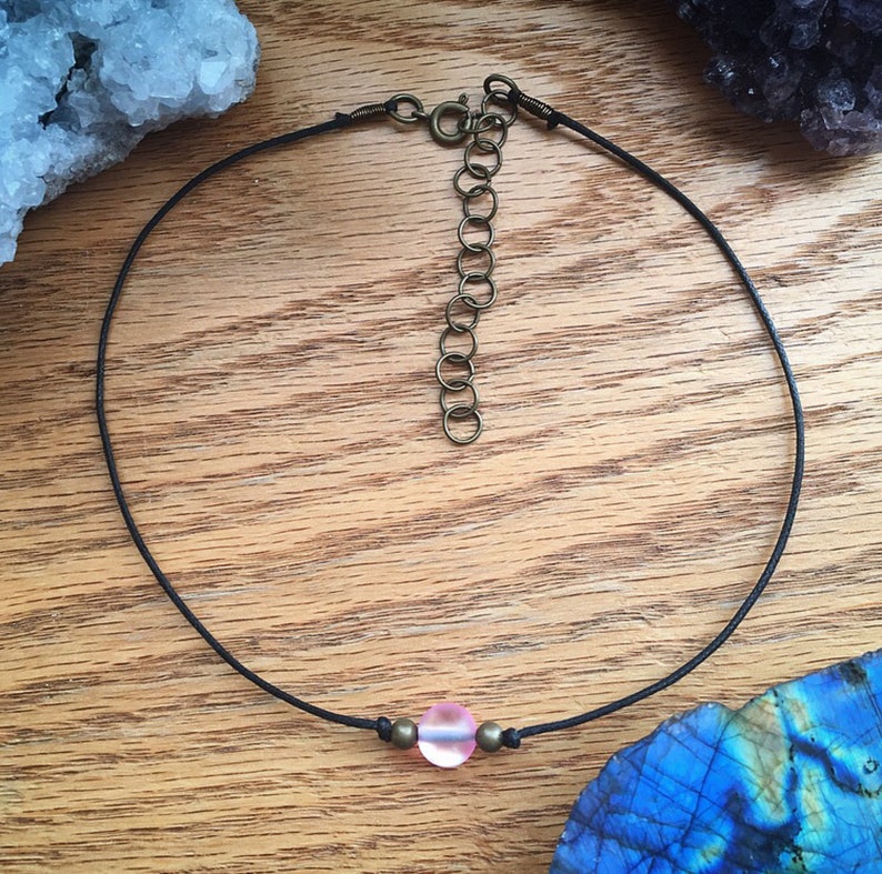 Puede incluir: Un delicado collar gargantilla con un cord&oacute;n negro y una cuenta central rosa de vidrio esmerilado. El collar tiene detalles met&aacute;licos de color lat&oacute;n antiguo y una cadena ajustable. Una joya sencilla y elegante.