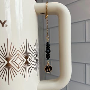 May include: A gold chain with black beads and a gold charm with the letter "A" on it. The charm is hanging from a white ceramic mug with a geometric pattern.