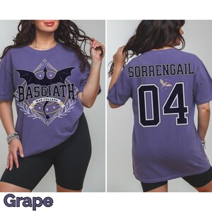 K&ouml;nnte beinhalten: Traubenfarbene T-Shirts mit grafischen Designs. Die Vorderseite zeigt ein schwarzes Drachendesign mit dem Text "BASGIATH WAR COLLEGE". Auf der R&uuml;ckseite steht "SORRENGAIL" &uuml;ber der Nummer "04".