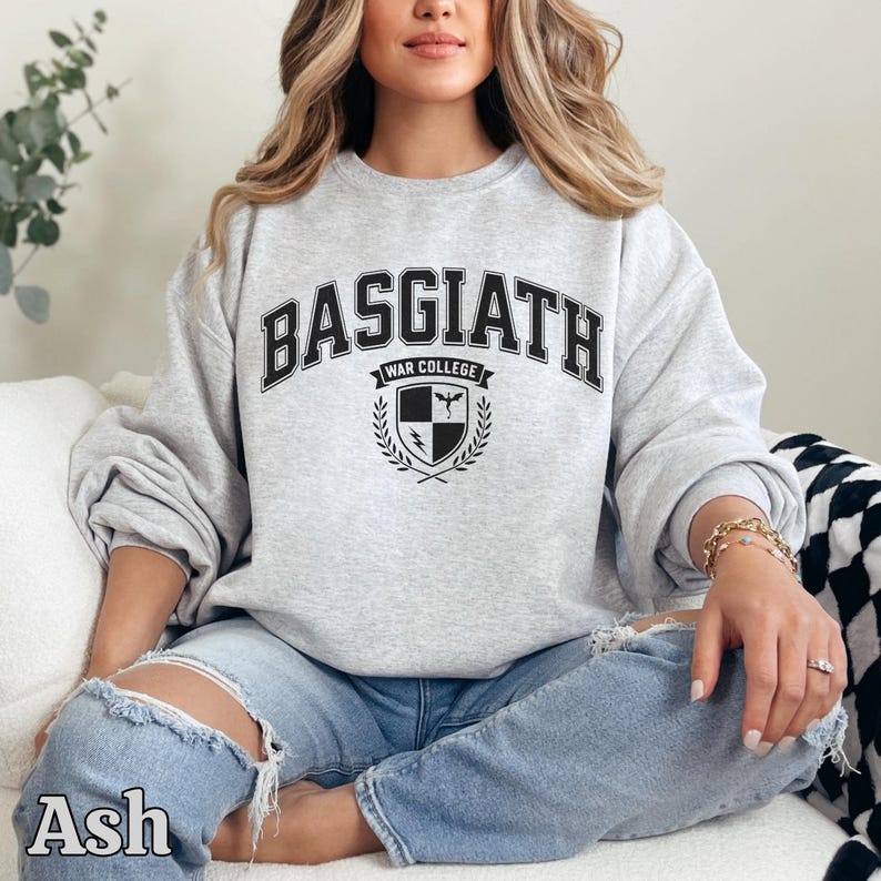 Peut inclure: Sweat-shirt gris clair avec l'inscription "BASGIATH WAR COLLEGE" en lettres noires. En dessous, un &eacute;cusson avec un bouclier, une &eacute;p&eacute;e et des branches de laurier. Le mot "Ash" est imprim&eacute; en bas &agrave; gauche.