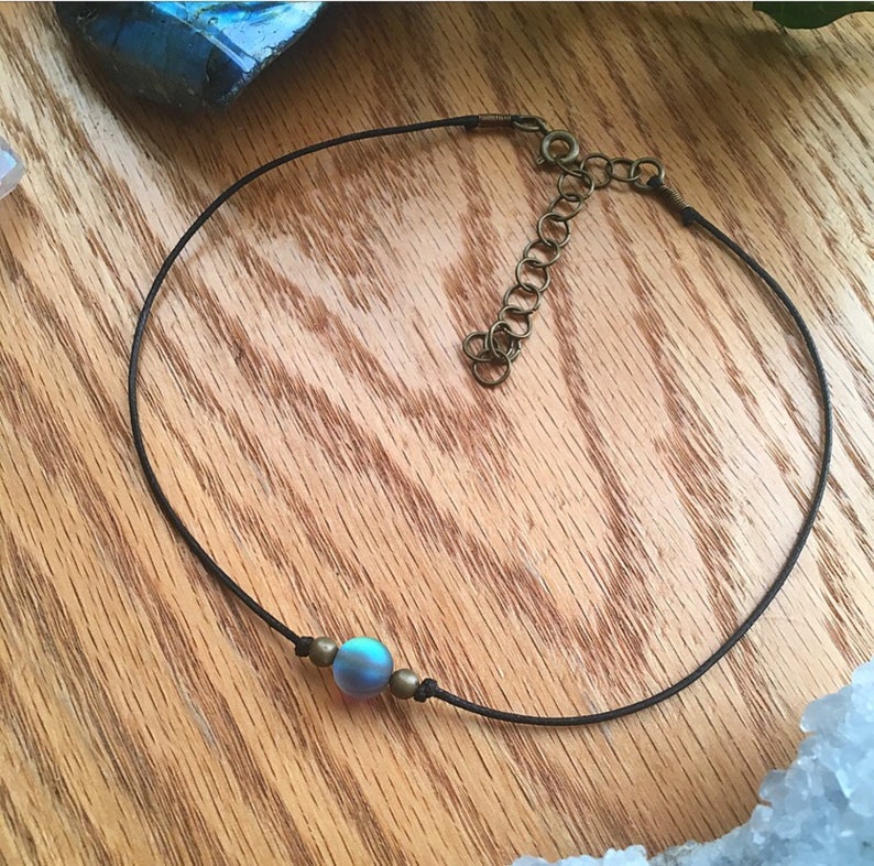 Puede incluir: Un collar gargantilla negro con una cuenta iridiscente azul-verde y detalles de lat&oacute;n antiguo. El collar presenta un cord&oacute;n fino y un cierre de cadena ajustable. La cuenta es el punto focal, agregando un toque de color y brillo al dise&ntilde;o minimalista.