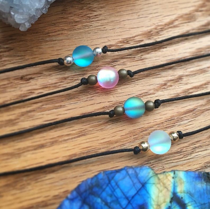 Puede incluir: Cuatro pulseras minimalistas con cuentas de vidrio iridiscente en tonos azules, rosas y verdes. Cada cuenta est&aacute; ensartada en un cord&oacute;n negro y acentuada con peque&ntilde;as cuentas de metal. Las pulseras se exhiben sobre una superficie de madera.