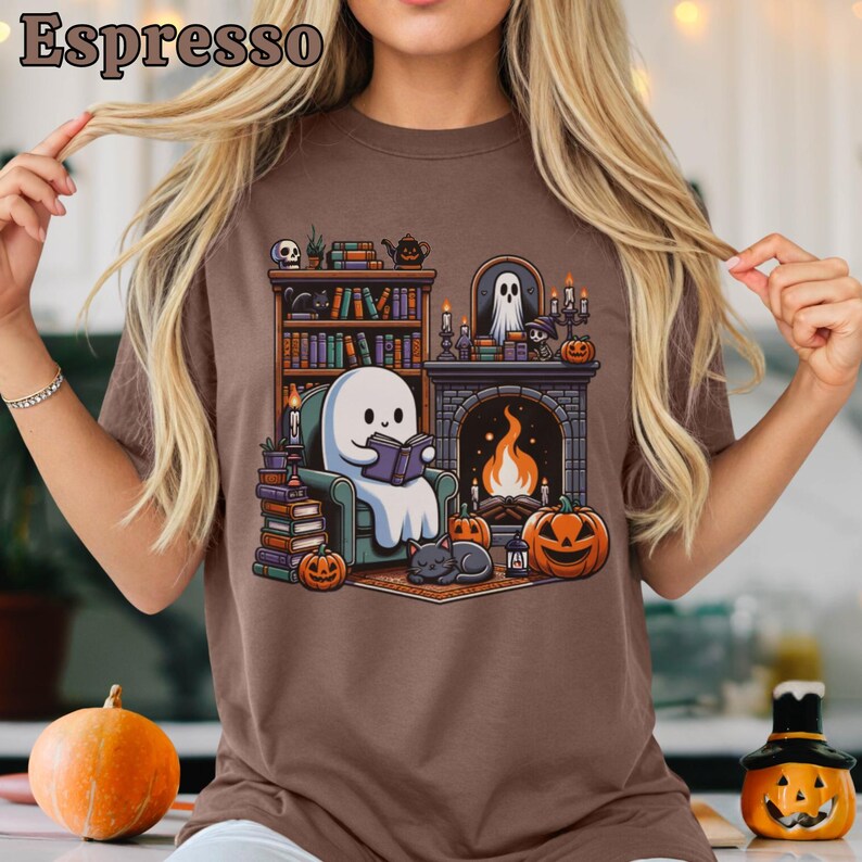 May include: Espresso-colored t-shirt featuring a Halloween-themed graphic. The design includes a cartoon ghost reading a book in a cozy setting with a fireplace, pumpkins, and a cat. The shirt is a comfortable, casual wear.