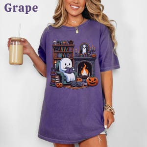 May include: A purple t-shirt with a graphic of a ghost reading a book in front of a fireplace, surrounded by pumpkins and a black cat. The word "Grape" is in the upper left corner. The shirt is a relaxed fit.