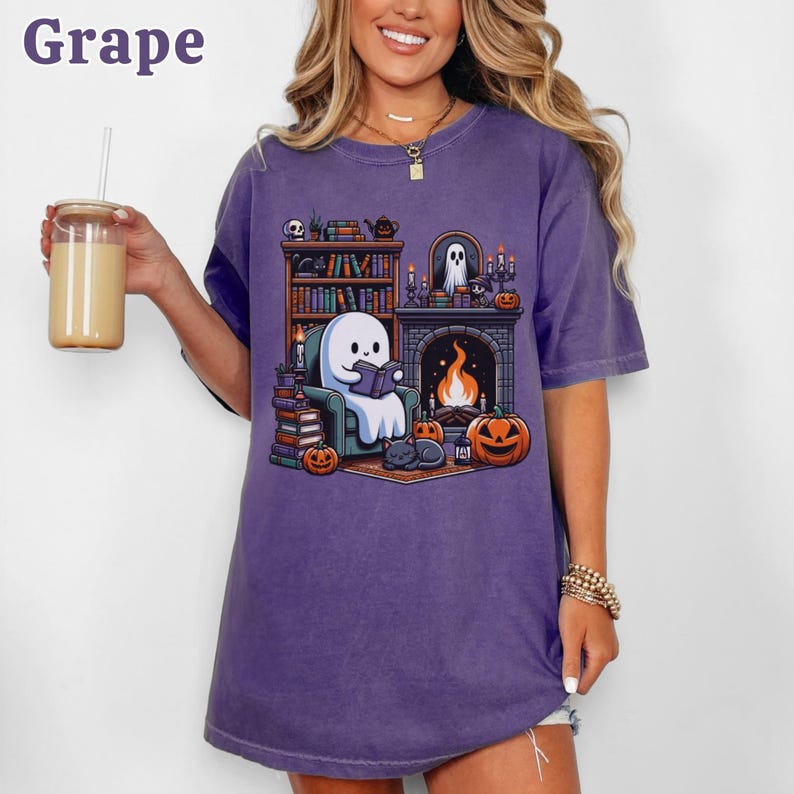 May include: A purple t-shirt with a graphic of a ghost reading a book in front of a fireplace, surrounded by pumpkins and a black cat. The word "Grape" is in the upper left corner. The shirt is a relaxed fit.