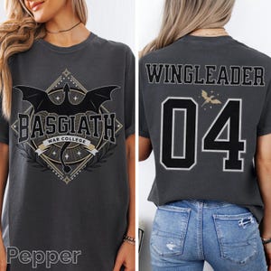 Op de afbeelding: Donkergrijs t-shirt met een drakenafbeelding en de tekst "BASGIATH WAR COLLEGE" op de voorkant. De achterkant van het shirt toont "WINGLEADER" en het nummer "04" in zwart en wit. Het shirt heeft een comfortabele pasvorm.