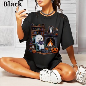 May include: Black t-shirt featuring a Halloween-themed graphic. The design includes a ghost reading a book by a fireplace, pumpkins, a cat, and a bookshelf. The word "Black" is printed at the top left.