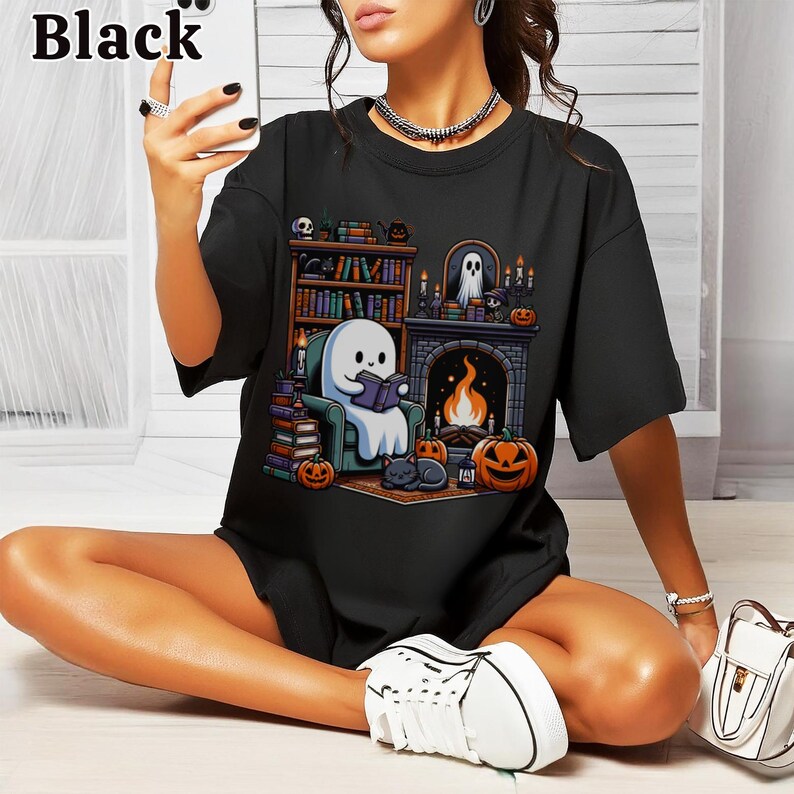 May include: Black t-shirt featuring a Halloween-themed graphic. The design includes a ghost reading a book by a fireplace, pumpkins, a cat, and a bookshelf. The word "Black" is printed at the top left.
