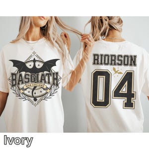 K&ouml;nnte beinhalten: Elfenbeinfarbene T-Shirts mit grafischen Designs. Eines zeigt "BASGIATH WAR COLLEGE" mit einem Drachenemblem. Das andere zeigt "RIORSON 04" mit einer kleinen Vogelgrafik. Die Shirts haben einen Rundhalsausschnitt und kurze &Auml;rmel.
