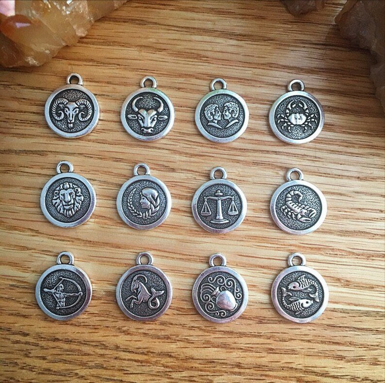 May include: Twelve silver charms featuring the astrological signs of the zodiac. The charms are round and have a raised design. The signs are: Aries, Taurus, Gemini, Cancer, Leo, Virgo, Libra, Scorpio, Sagittarius, Capricorn, Aquarius, and Pisces.