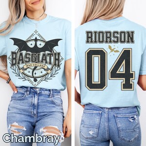 K&ouml;nnte beinhalten: Hellblaues T-Shirt mit einem Aufdruck auf der Vorderseite, der einen schwarzen Drachen, den Text "BASGIATH WAR COLLEGE" und einen dekorativen Rahmen zeigt. Auf der R&uuml;ckseite steht "RIORSON 04" in Schwarz und Gold. Das Wort "Chambray" steht unten.