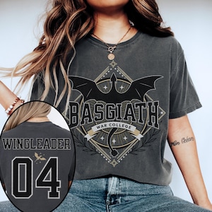 Vintage-style Basgiath War College tee inspired by Fourth Wing. Front features bold collegiate lettering with a bat-winged crest and star accents. Back shows customizable jersey-style design with ‘WINGLEADER’ and the number 04, plus a subtle golden dragon silhouette. Printed on a washed black Comfort Colors-style shirt for a soft, lived-in look. Perfect for fantasy fans and Riders Quadrant enthusiasts.