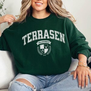 以下が含まれることがあります： 白い文字で「TERRASEN」と書かれた濃い緑色のスウェットシャツ。「GALATHYNIUS」と鹿と盾の紋章が下にあります。スウェットシャツはクルーネックです。サイズはセンチメートルです。