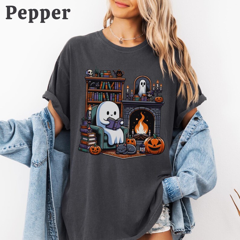 May include: Dark gray t-shirt featuring a cartoon ghost reading a book in front of a fireplace. The scene includes a bookshelf, pumpkins, a black cat, and the word "Pepper" at the top left.