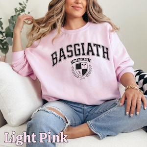Peut inclure: Sweat-shirt rose clair avec l'inscription "BASGIATH WAR COLLEGE" en noir. Le design comprend un &eacute;cusson avec un bouclier, un oiseau et un &eacute;clair. Les mots "Light Pink" sont &eacute;crits en cursive en bas.